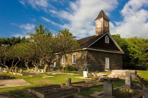 A stone church with a bell tower surrounded by trees and a cemetery under a blue sky with scattered clouds.