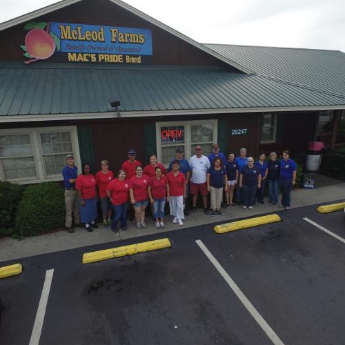 A group of employees stands outside McLeod Farms, a farm store, with a sign indicating it's open.