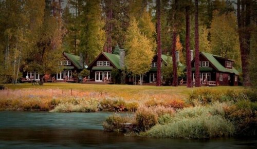 A serene lakeside scene featuring three rustic cabins surrounded by tall trees and colorful autumn foliage.