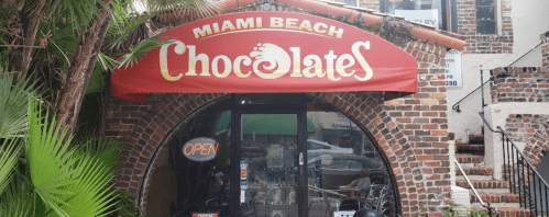 A storefront with a red awning reading "Miami Beach Chocolates," surrounded by greenery and brick architecture.