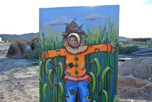 A child peeks through a cutout of a colorful scarecrow in a cornfield setting.