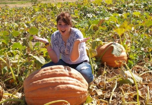 A person sitting among large pumpkins in a field, smiling and gesturing with hands raised in surprise.