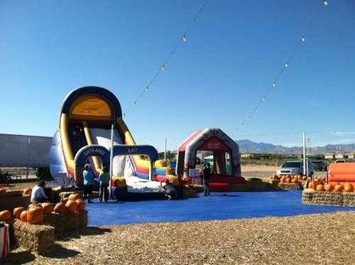 A colorful inflatable slide and bounce house at a pumpkin patch, surrounded by hay bales and pumpkins under string lights.