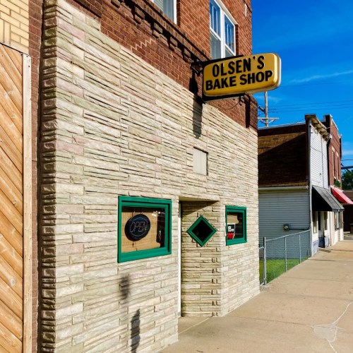 Exterior of Olsen's Bake Shop with a textured stone facade and a bright "OPEN" sign on a sunny day.