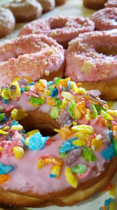 A close-up of two colorful donuts: one with pink frosting and sprinkles, and another topped with fruity cereal.