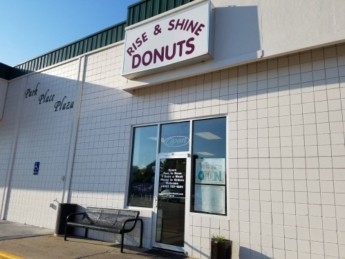 Exterior of "Rise & Shine Donuts" shop with a sign and open door, located in Park Place Plaza. Bench outside.