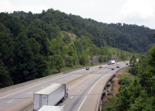 A highway with vehicles traveling, surrounded by lush green hills and trees under a cloudy sky.