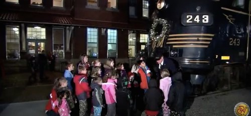 A group of children gathers around a decorated train, listening to an adult in front of a historic building.
