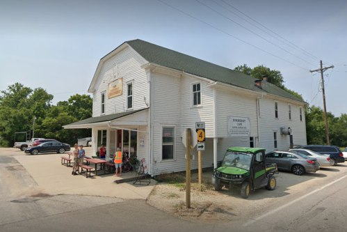 A white two-story building with a green roof, featuring outdoor seating and parked vehicles, set in a rural area.