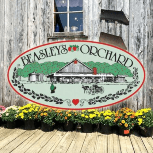 Sign for Beasley's Orchard with a rustic barn and flower pots in front, set against a weathered wooden background.