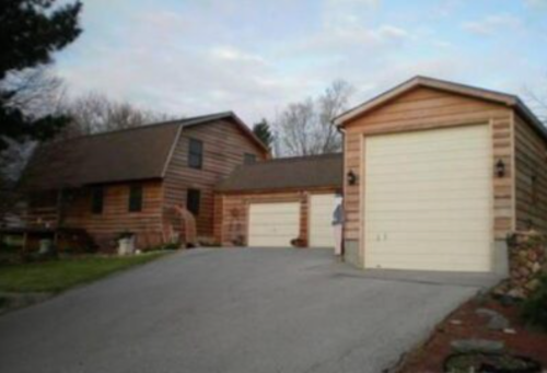 A wooden house with a sloped roof and a garage, set on a paved driveway surrounded by trees.
