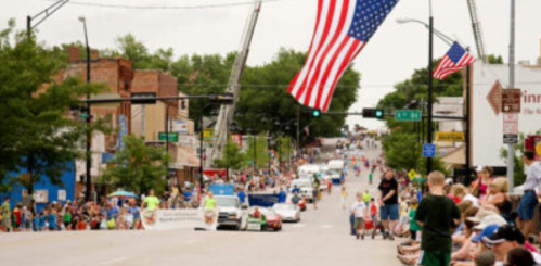 A lively parade scene with crowds lining the street, American flags, and vehicles moving down the road.