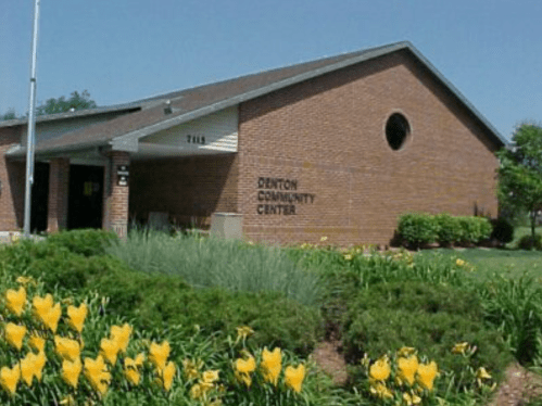 A brick building with a sign reading "Denton Community Center," surrounded by green grass and yellow flowers.
