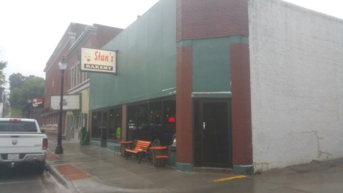 A bakery storefront with a green facade and a sign reading "Stan's Bakery," featuring outdoor seating and rainy weather.