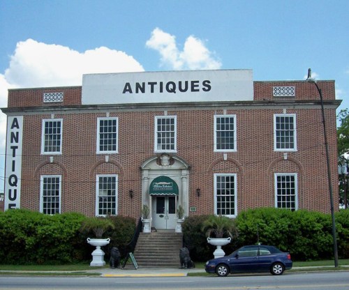 A brick building with large windows and a sign reading "ANTIQUE," surrounded by greenery and decorative planters.