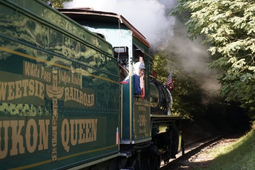 A vintage steam train, the Yukon Queen, travels along a scenic track with a conductor visible in the window.