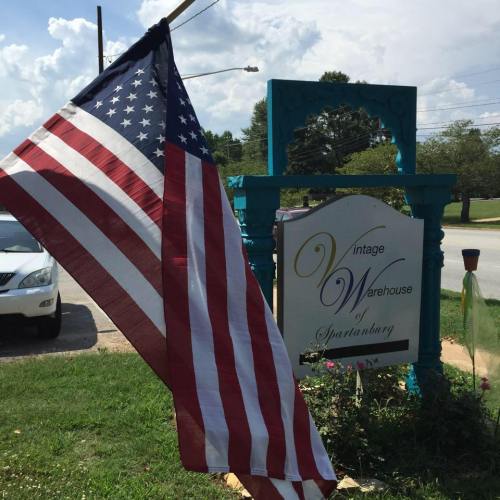 An American flag waves in front of a vintage warehouse sign in Spartanburg, surrounded by greenery and a clear sky.
