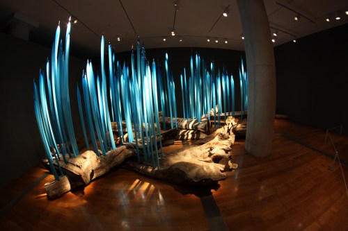 Art installation featuring a log with vibrant blue glass spikes resembling plants, set in a modern gallery space.