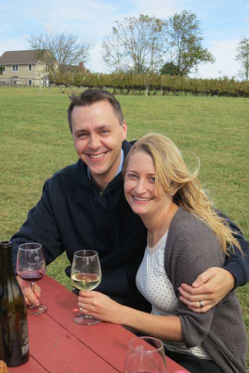 A smiling couple sits at a picnic table outdoors, holding wine glasses, with a vineyard and blue sky in the background.