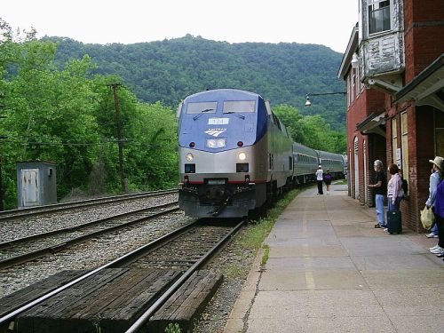 A train approaches a small station surrounded by green hills, with passengers waiting on the platform.