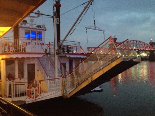 A riverboat docked at twilight, with a bridge illuminated in the background and a lifebuoy visible on the side.