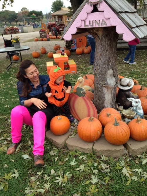 A woman and a child in a pumpkin patch, surrounded by pumpkins and autumn decorations, smiling and enjoying the day.