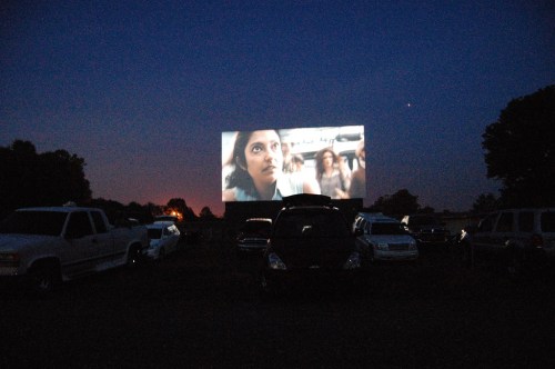 A drive-in movie screen shows a scene at dusk, surrounded by parked cars in a field.