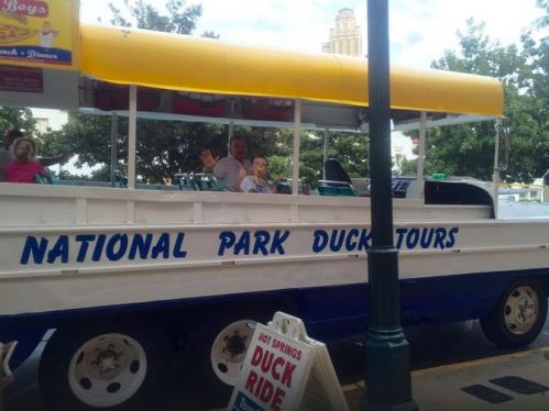 A yellow and blue duck boat for tours, with passengers waving, parked in a city setting.