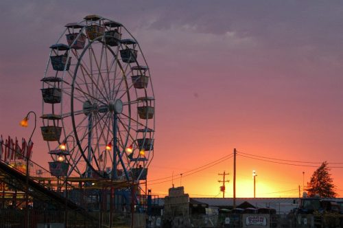 A ferris wheel silhouetted against a vibrant sunset with colorful clouds and lights in the foreground.