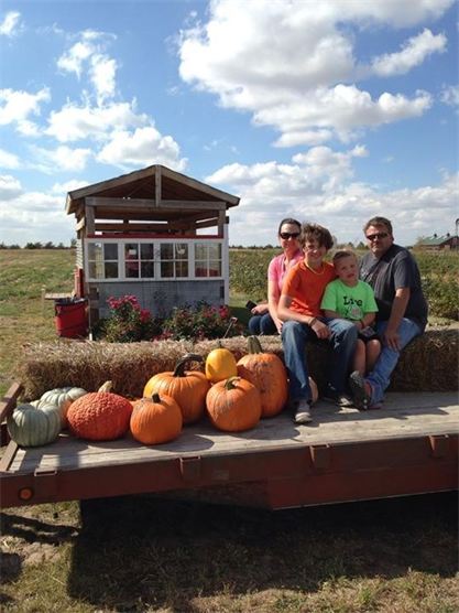 A family sits on a hay wagon with pumpkins, in front of a small shed and a field under a blue sky with clouds.