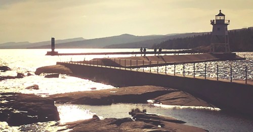 A lighthouse stands on a pier extending into a shimmering body of water, with rocky shores and distant hills in the background.