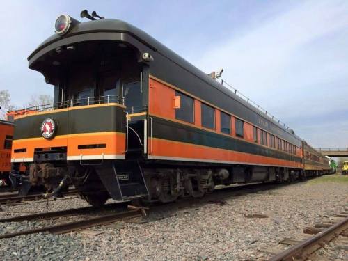 A vintage train car with a black and orange exterior, parked on gravel tracks under a clear sky.