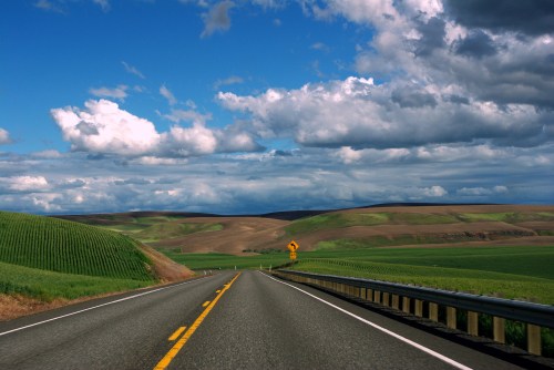 A winding road stretches through green fields under a blue sky with fluffy clouds.