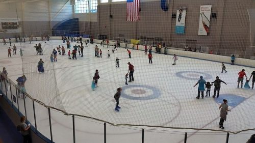A busy ice skating rink with many people skating and enjoying the activity, surrounded by a netted barrier.