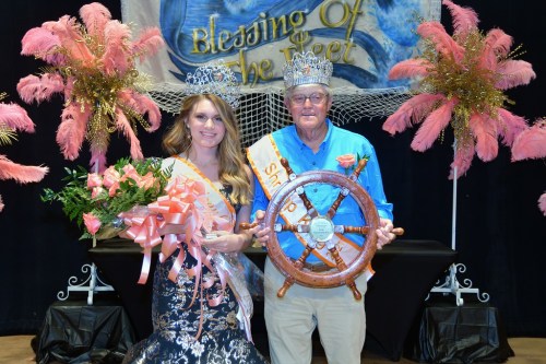 A woman in a crown and gown holds flowers next to a man in a blue shirt, both smiling at a festive event.