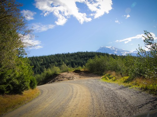 A winding dirt road surrounded by greenery, leading towards a mountain under a blue sky with scattered clouds.
