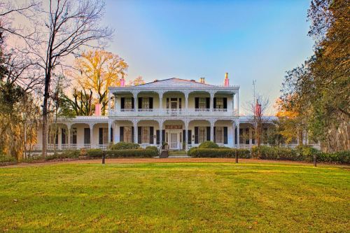 A grand, historic mansion with a wraparound porch, surrounded by lush greenery and trees under a clear blue sky.