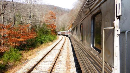 A train curves along a track surrounded by autumn trees and mountains in the background.