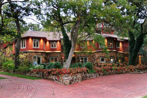 A charming two-story house with stone and wood exterior, surrounded by lush greenery and colorful plants.
