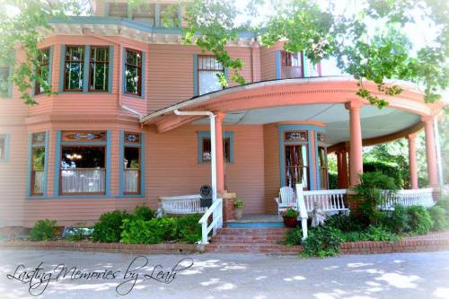 A charming pink house with a wraparound porch, surrounded by greenery and flower pots.