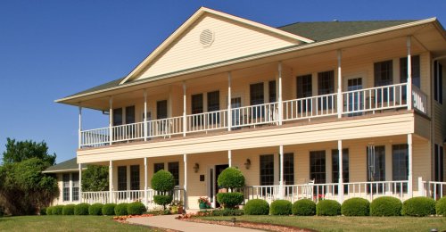 A large, two-story house with a wraparound porch, green roof, and neatly trimmed bushes in front under a clear blue sky.