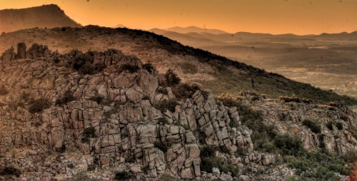 Sunset over rugged mountains, showcasing rocky terrain and distant hills under a warm orange sky.