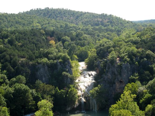 A scenic view of a waterfall cascading down rocky cliffs, surrounded by lush green trees and hills in the background.