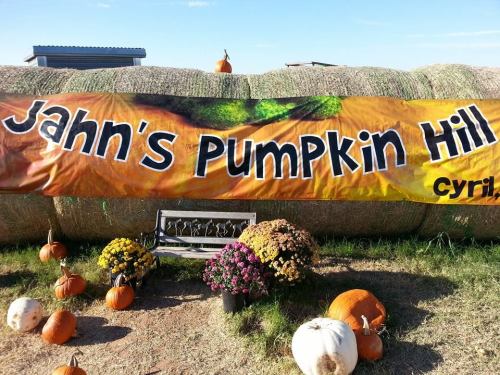 A banner reading "Jahn's Pumpkin Hill" with pumpkins and flowers in front of hay bales under a clear blue sky.