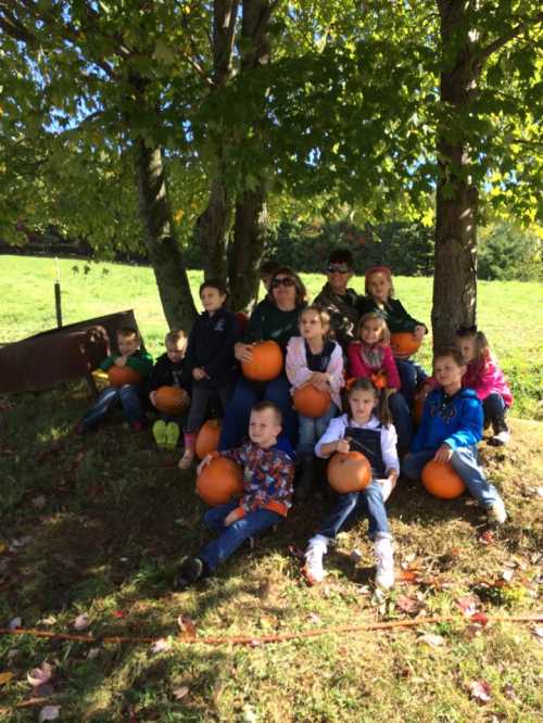 A group of children and adults sitting under trees, holding pumpkins, with a grassy field in the background.