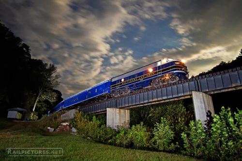 A blue vintage train crosses a bridge under a cloudy night sky, surrounded by greenery.