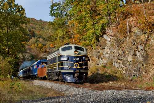 A vintage train travels along a scenic track surrounded by autumn foliage and rocky hills.