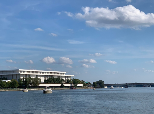 A calm river scene with boats and a modern building under a blue sky with fluffy clouds.