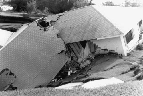 A house partially collapsed, with a damaged roof and exposed interior, surrounded by uneven ground.