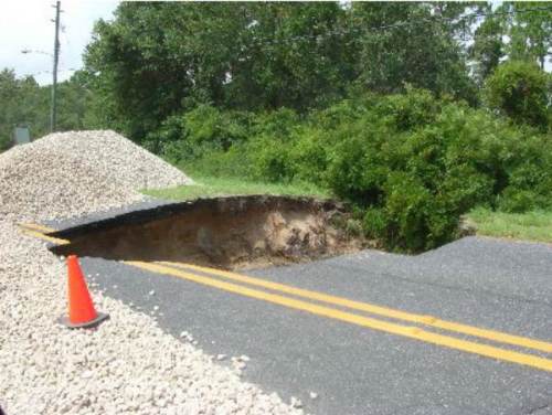 A road with a large sinkhole, surrounded by gravel and a traffic cone, with greenery in the background.